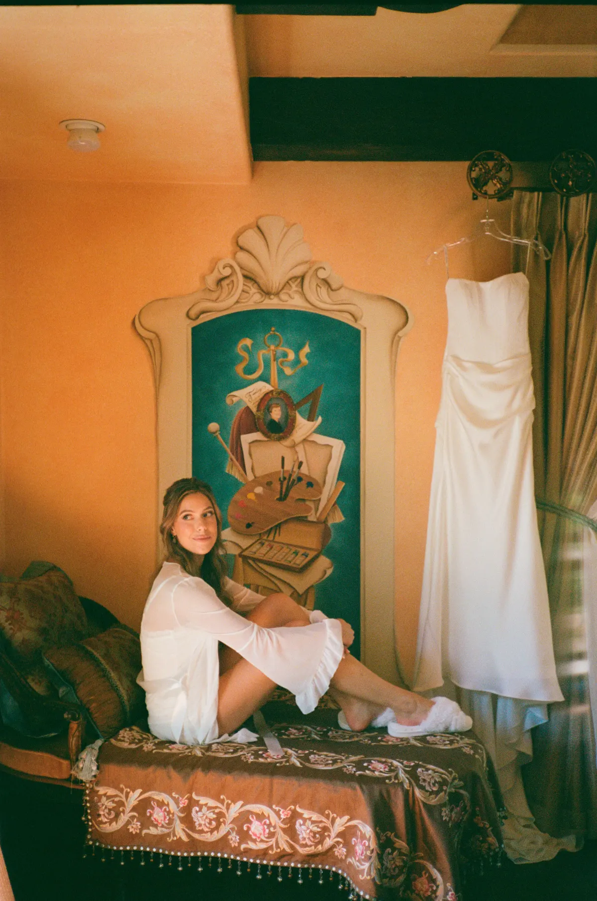 Bride on ornate sofa, wedding dress hanging beside her