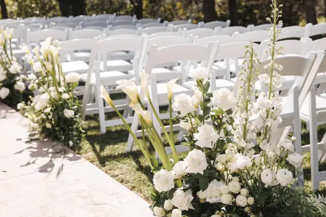White floral arrangements line the aisle beside rows of white folding chairs set up for an outdoor wedding ceremony.