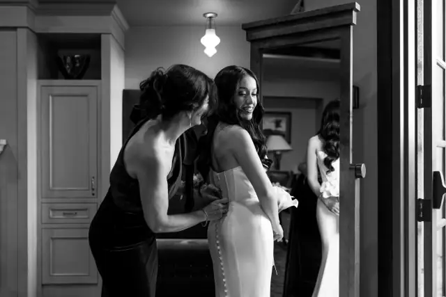 Bride Grace smiling while a woman helps button her wedding dress in a cozy indoor room.