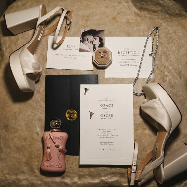 Flat lay of wedding details including Grace and Oscar's invitations, RSVP card, engagement ring in velvet box, pair of ivory platform heels, perfume bottle, and diamond necklace on a tan textured surface.
