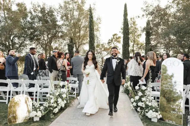 Bride and groom holding hands and smiling as they walk down the aisle after their outdoor wedding ceremony surrounded by guests and floral decorations.