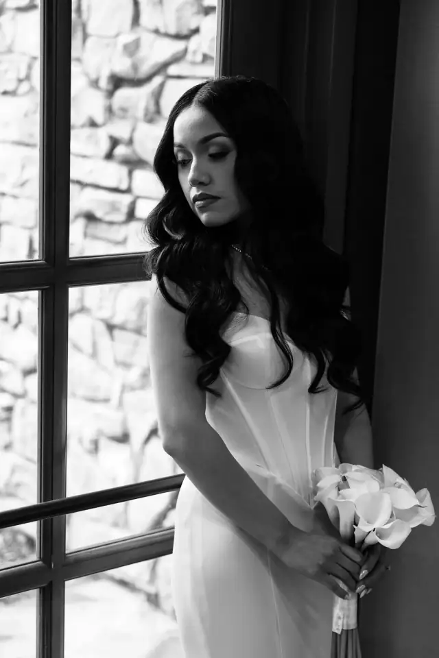 Black and white photo of a bride in a fitted wedding dress holding a bouquet of calla lilies while standing by a window with a stone wall outside.