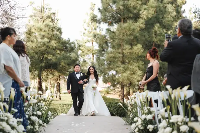 Bride in white wedding dress walking arm-in-arm with man in tuxedo down floral aisle outdoors surrounded by guests at Grace & Oscar's wedding
