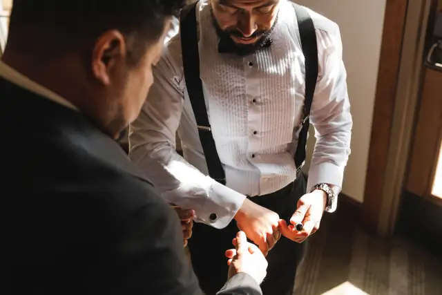 Groom and groomsman adjusting cufflinks indoors in natural light during wedding preparations.