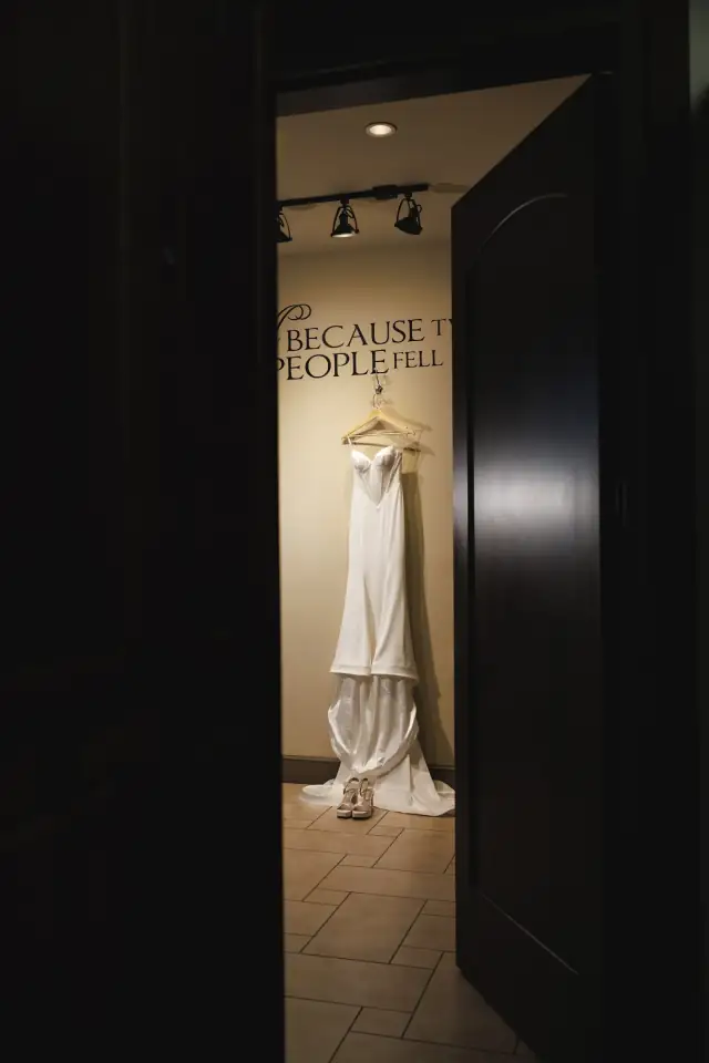 Wedding dress hanging on a wall with high heels beneath, viewed through a partially open dark door in a softly lit room.