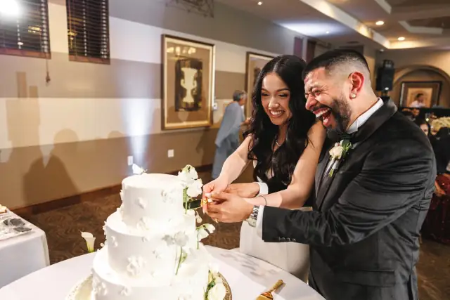 Grace and Oscar joyfully cut their three-tier white wedding cake indoors during their reception.