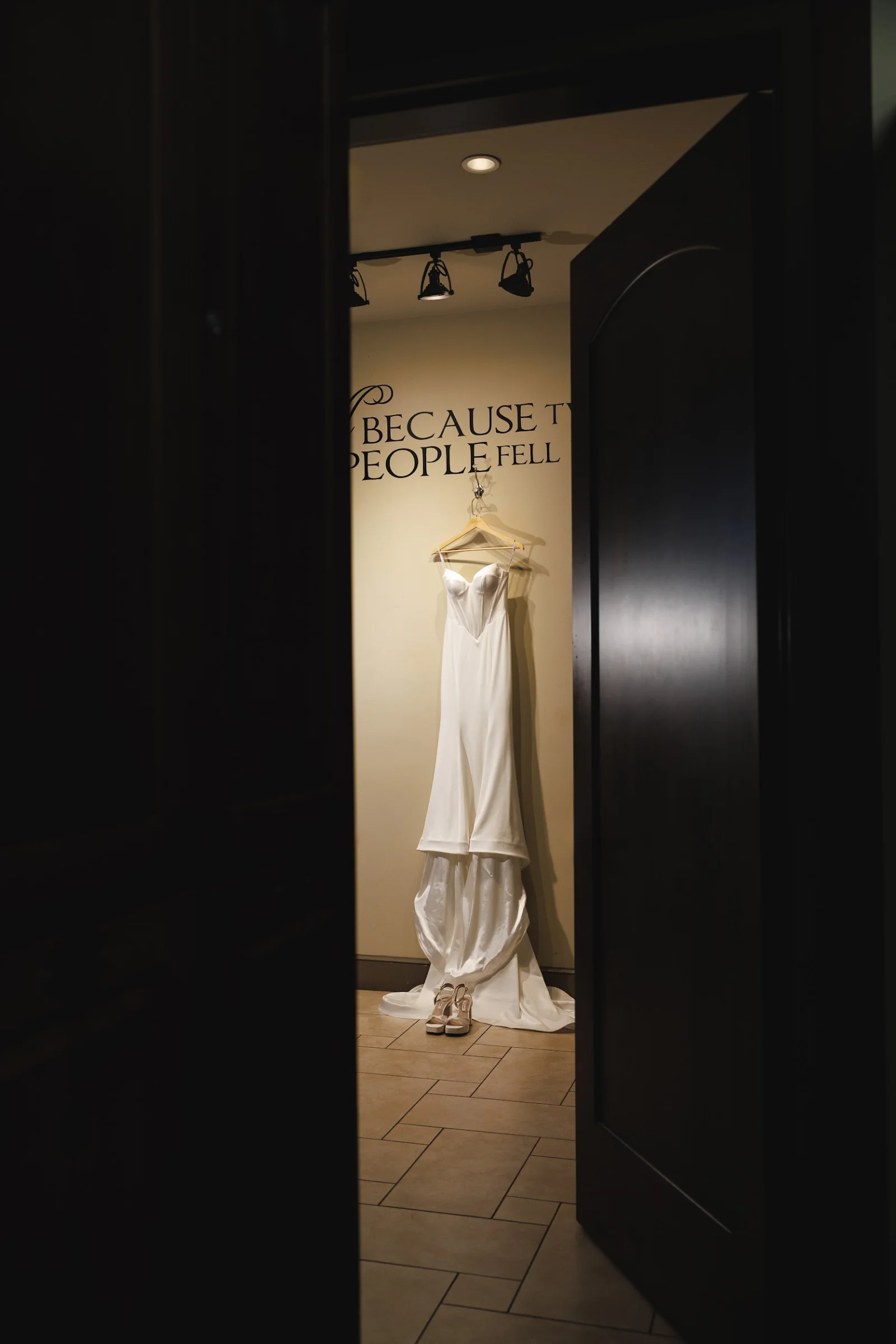 Wedding dress hanging on a wall with high heels beneath, viewed through a partially open dark door in a softly lit room.