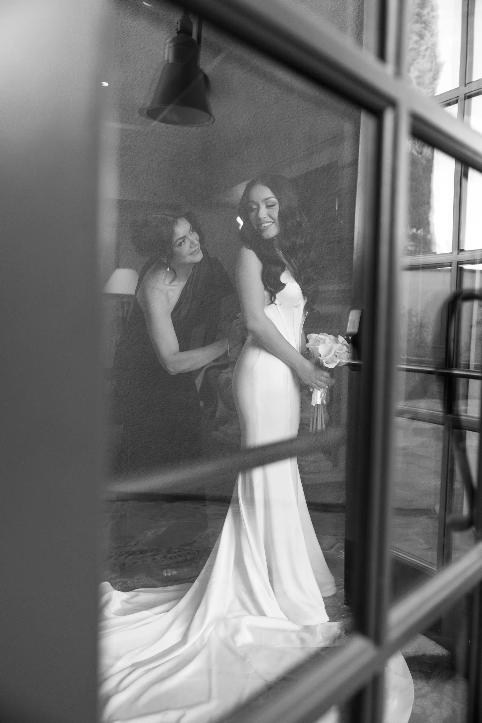 Black and white photo of a bride in a wedding gown holding a bouquet while a bridesmaid adjusts her dress indoors, seen through a window.