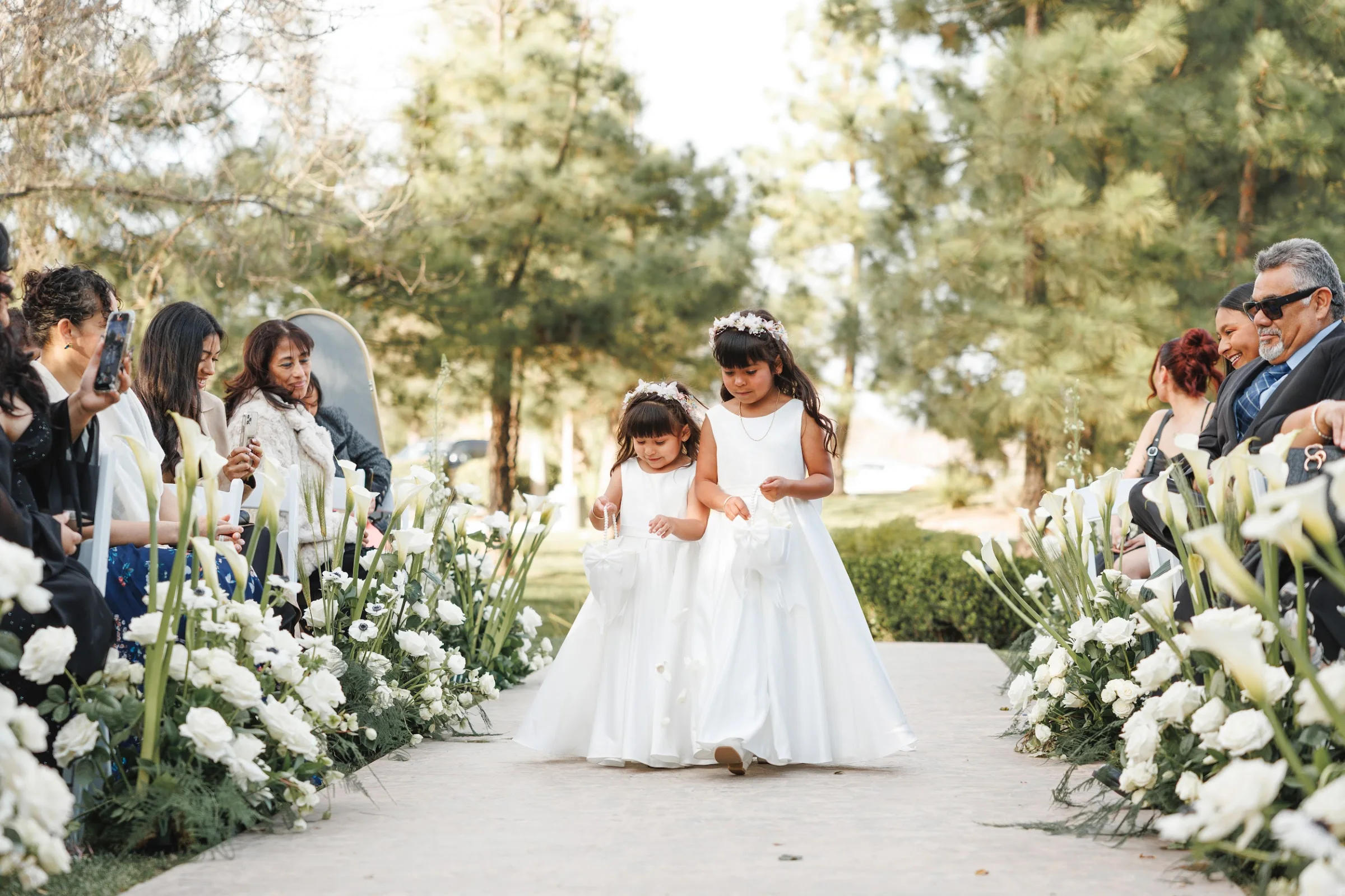 Two young flower girls in white dresses walking down a flower-lined aisle outdoors during a wedding ceremony.