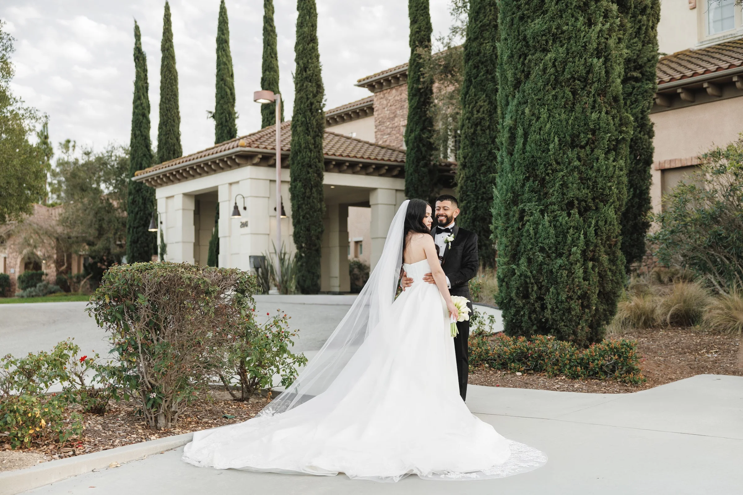 Bride in white gown and veil holding bouquet embraced by groom in tuxedo outside a Mediterranean-style building with tall cypress trees.