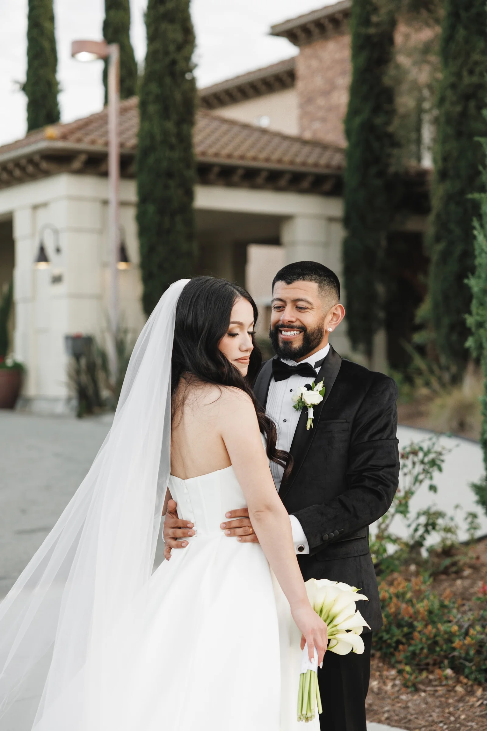 Bride in white gown holding bouquet and groom in black tuxedo smiling at each other outdoors with tall trees and building in background.