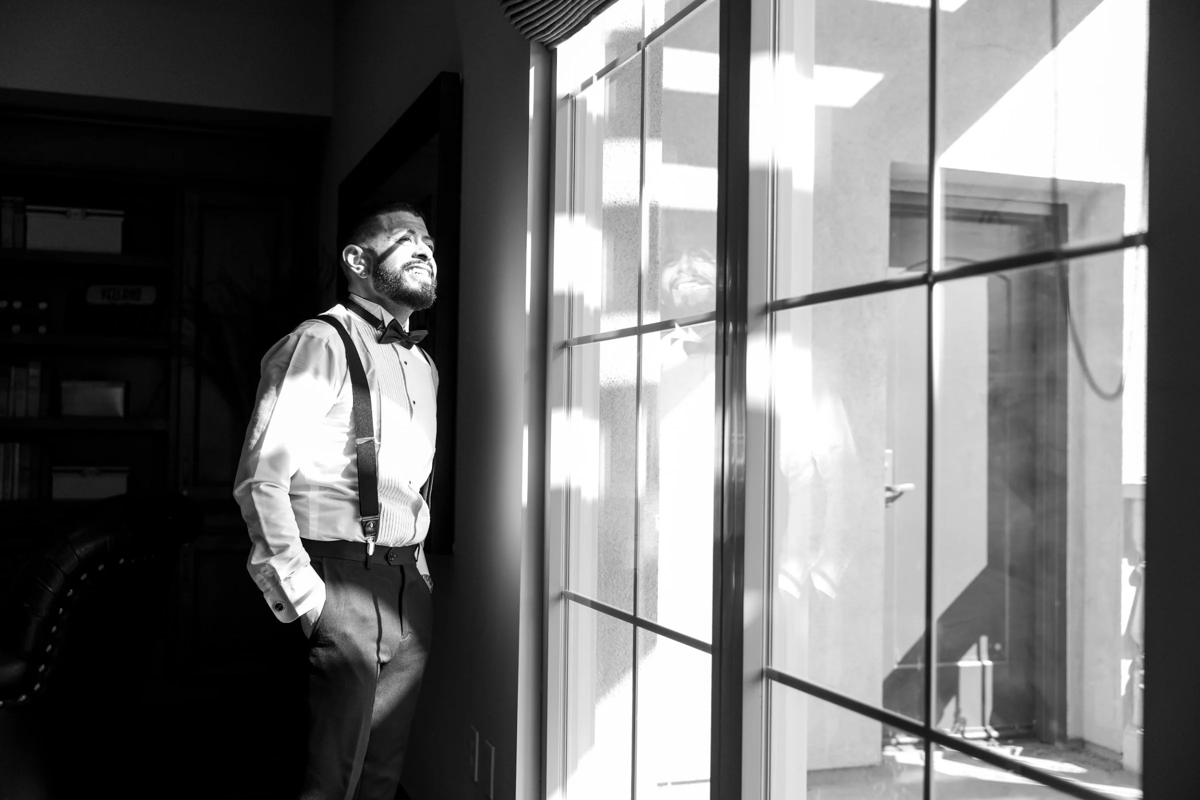 Groom in tuxedo with suspenders standing indoors by a window with sunlight and reflection, captured in a black and white wedding photo.