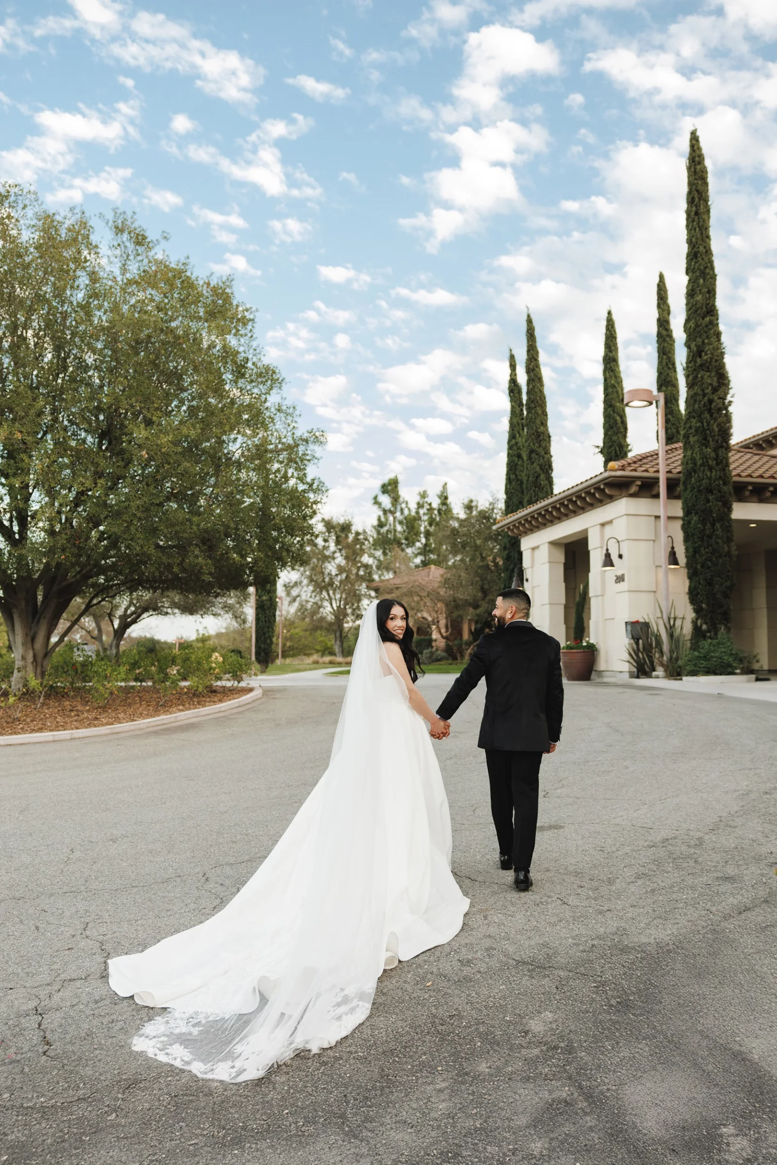 Bride in a flowing white wedding dress and veil holding hands with groom in a black suit, walking outdoors near a building with tall cypress trees under a partly cloudy sky.
