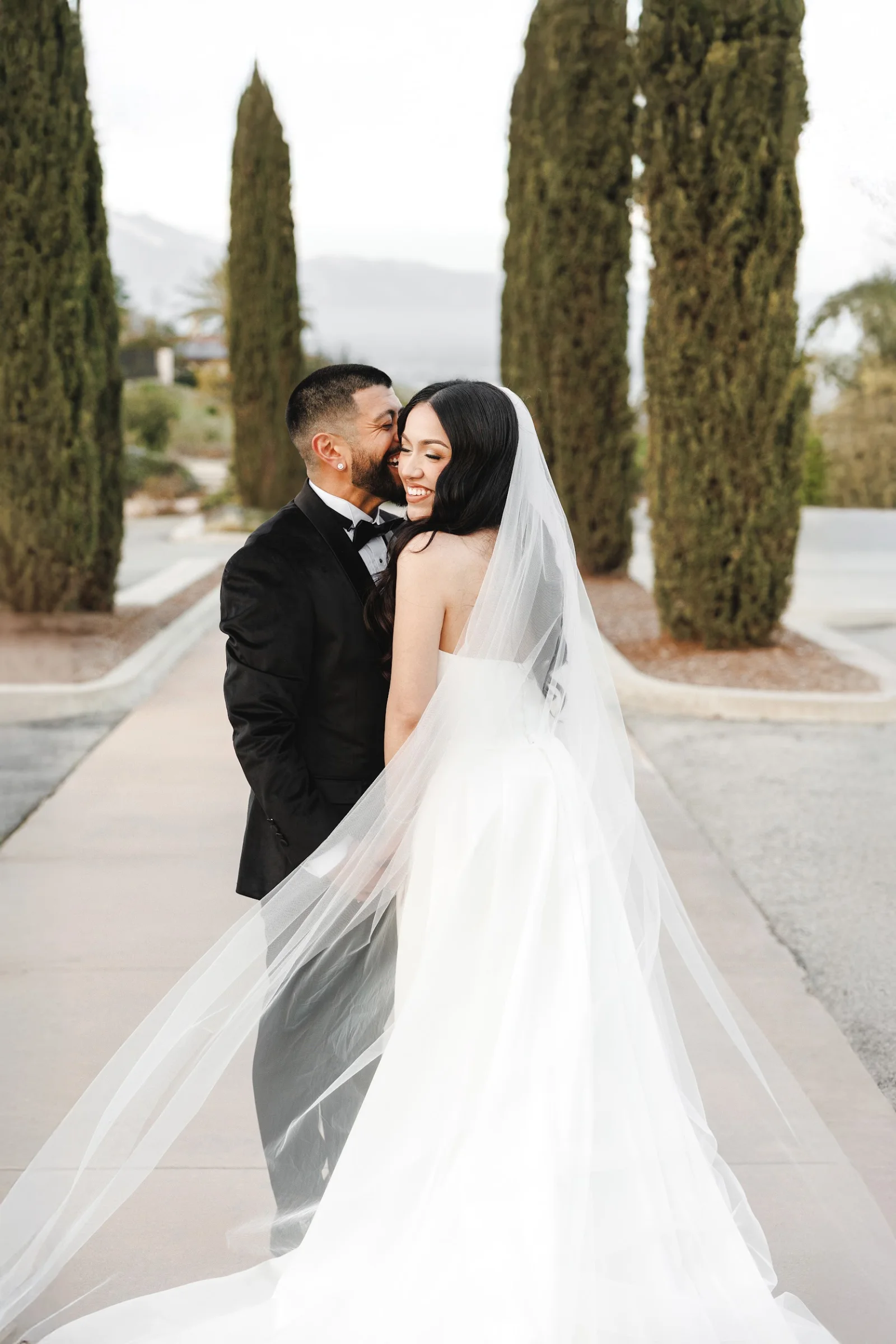 Bride in white dress and long veil smiling with groom in black tuxedo on outdoor path lined with tall trees.