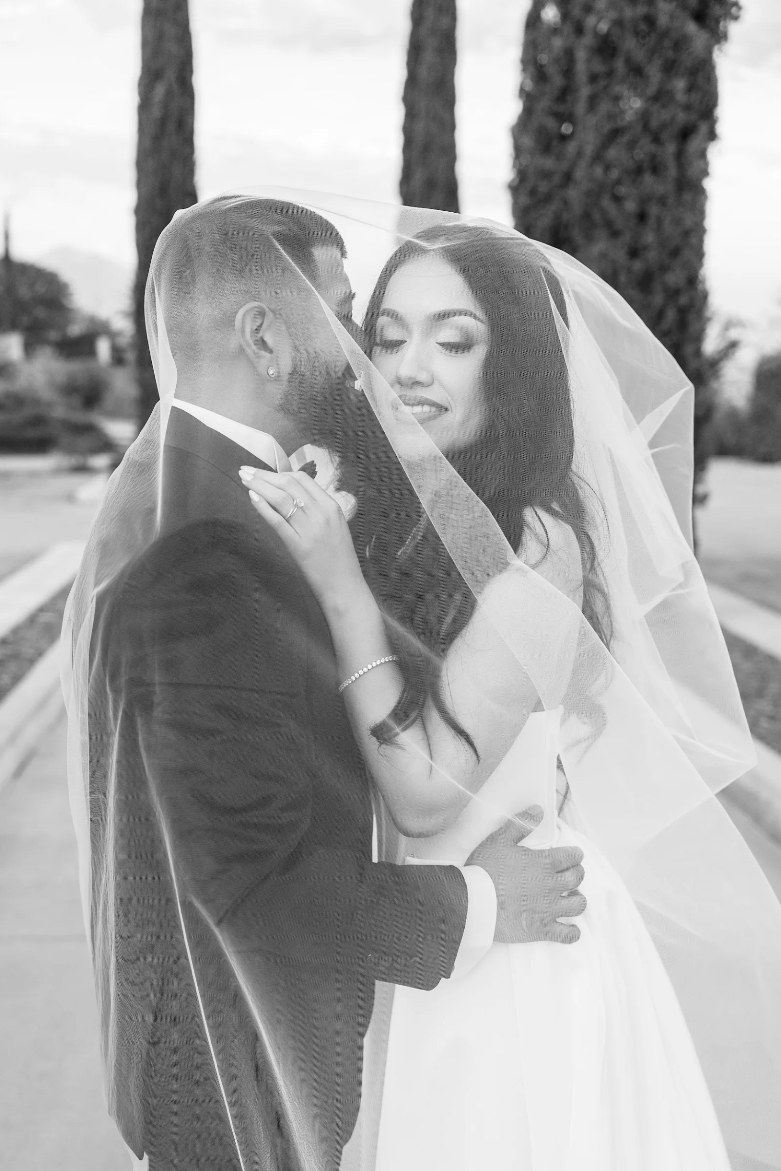 Bride and groom embrace under a wedding veil outdoors with tall trees in the background.