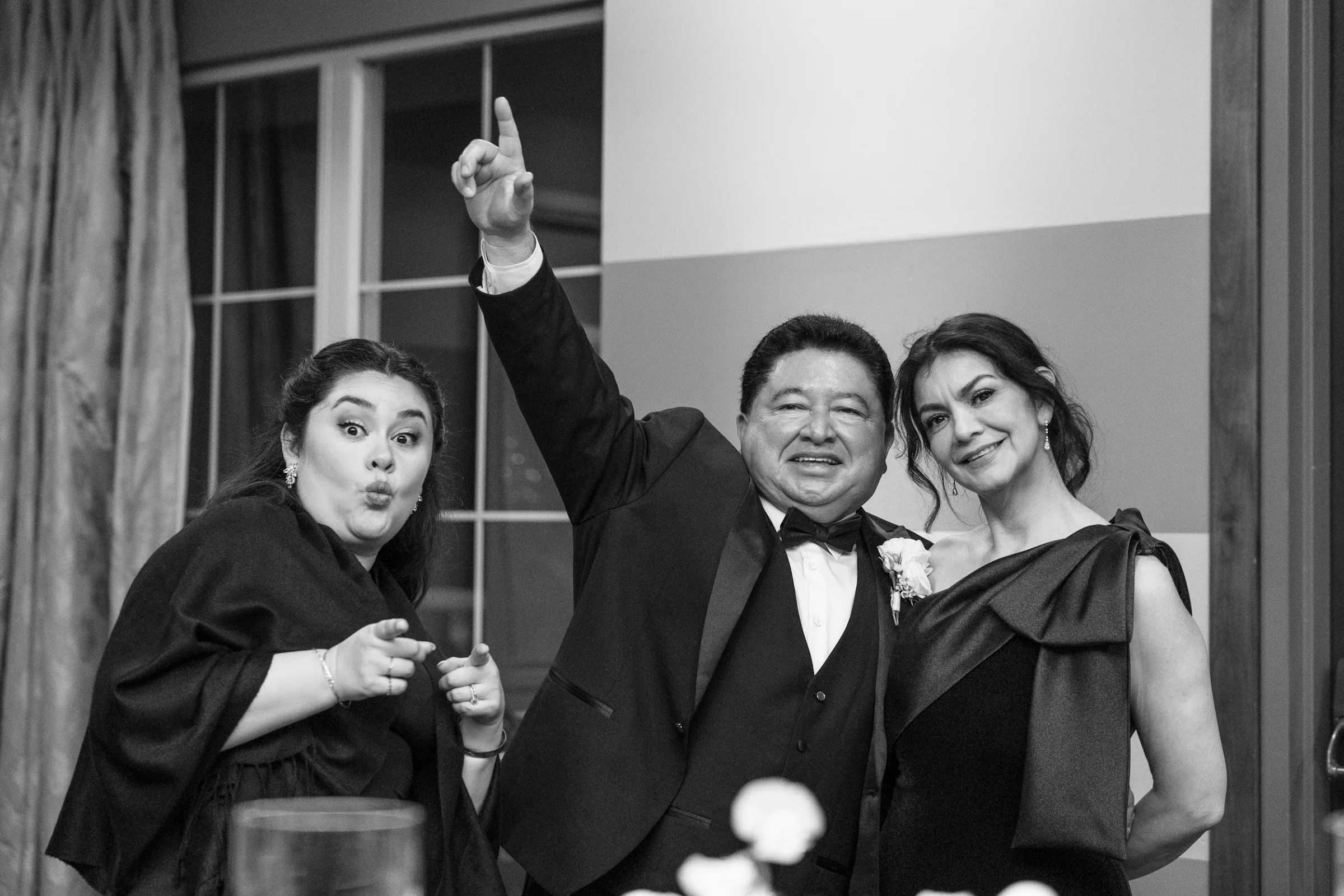 Black and white photo of three wedding guests posing happily indoors, with a man in a tuxedo and two women in elegant dresses at a formal event.